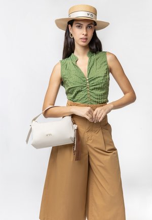 Mujer con blusa verde sin mangas, pantalones anchos color tostado, sombrero de paja, sosteniendo un bolso blanco con correa marrón sobre un fondo liso.