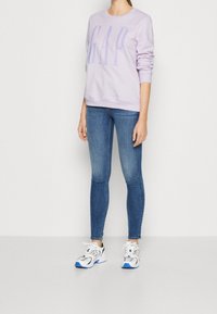 Felpa color lavanda con logo "GAP", abbinata a jeans aderenti blu e scarpe da ginnastica bianche con dettagli neri e blu. Tessuto piatto, design casual.