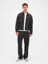 Homem vestindo um fato preto riscado e calças combinando com uma camisa branca e ténis pretos, posando contra um fundo branco liso.