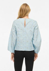 Femme blonde portant un chemisier bleu clair à manches longues avec des motifs floraux en découpe, et un pantalon noir ajusté, vue de dos.