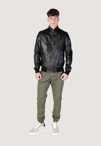 Giubbotto bomber in pelle nera con polsini e colletto a costine, chiusura con zip e dettagli del logo. Abbinato a pantaloni verde oliva e sneakers grigie.