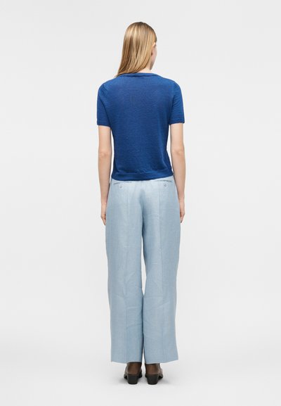 Γυναίκα φαινομένη από πίσω, ντυμένη με μπλε μπλούζα με κοντά μανίκια, ανοιχτό μπλε παντελόνι φαρδύ στις μπατζάκια και μαύρες ψηλοτάκουνες γόβες, μπροστά σε λευκό, απλό φόντο.