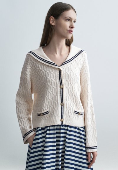 Femme portant un cardigan crème en maille torsadée avec des bordures bleu marine et des boutons dorés, associé à une jupe à rayures horizontales bleues et blanches.