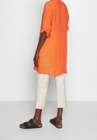 Tunika in lino arancione brillante con maniche arrotolate, abbinata a pantaloni cropped beige chiaro e sandali neri con due cinturini e suole in sughero.