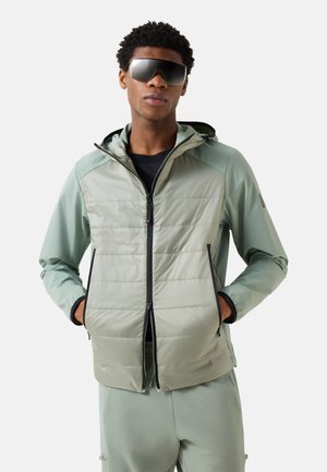 Mand iført lysegrøn hættetrøje med sort lynlås og matchende bukser, hænderne i lommerne, iført store sorte solbriller, neutralt baggrund.