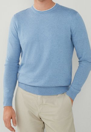 Uomo che indossa un maglione azzurro chiaro con scollo rotondo e pantaloni beige, una mano in tasca su sfondo semplice.