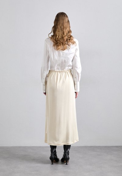 Camicia bianca con un design strutturato, abbinata a una gonna crema di lunghezza al ginocchio con vita elasticizzata e una leggera lucentezza, indossata con stivali neri.