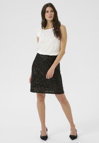 Top blanco sin mangas con detalle de lazo; falda negra de lentejuelas hasta la rodilla; modelo con tacones negros de punta; fondo claro liso.