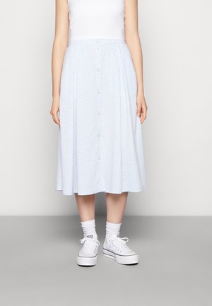 Personne portant un débardeur blanc, une jupe midi bleu clair boutonnée avec un motif subtil, des chaussettes blanches et des baskets à plateformes blanches.