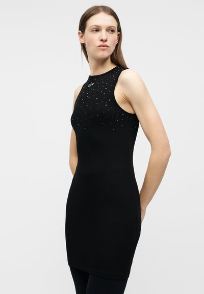 Γυναίκα που φοράει στενή μαύρη αμάνικη φορεμα με μικρές ασημένιες καρφίτσες και τη λέξη «OFF» κοντά στη γραμμή του λαιμού, στέκεται μπροστά σε απλό φόντο.