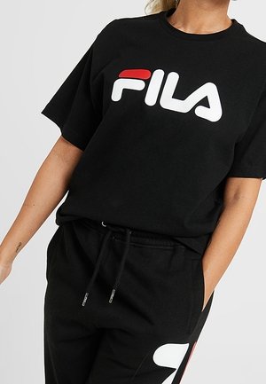 Person iført en sort FILA t-shirt og matchende sorte bukser med hvide og røde logoaccents, stående med den ene hånd i lommen.