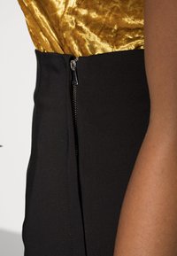 Schwarze Rock mit einem seitlichen Reißverschluss und metallischen Beschlägen. Glatte Textur mit einer taillierten Passform, kombiniert mit einem glänzenden goldenen Oberteil.