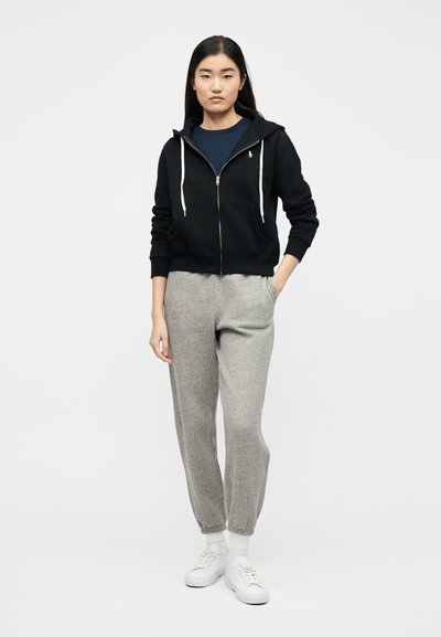 Γυναίκα στέκεται φορώντας μαύρο φούτερ με φερμουάρ, μπλε σκουρόχρωμο πουκάμισο, γκρι φόρμα, λευκές κάλτσες και λευκά αθλητικά παπούτσια μπροστά σε απλό λευκό φόντο.