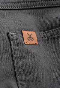 Tissu gris avec une surface texturée, comportant une étiquette en cuir marron embossée avec des ciseaux, cousue sur la poche.