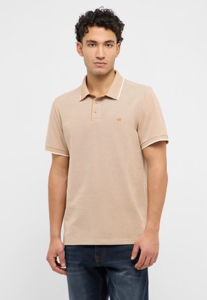 Junger Mann mit lockigem dunklem Haar, der ein beiges Poloshirt mit kurzen Ärmeln und dunkelblaue Jeans vor einem schlichten weißen Hintergrund trägt.