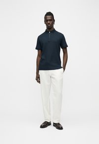 Homme debout devant un fond uni, portant un polo bleu marine à manches courtes et fermeture éclair, un pantalon blanc et des chaussures marron foncé.