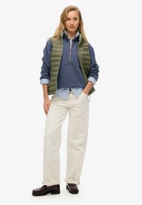 Grön puffig väst över en marinblå tröja med halvmatch, lager över en ljusblå randig skjorta. Vita vida byxor och mörka skor.