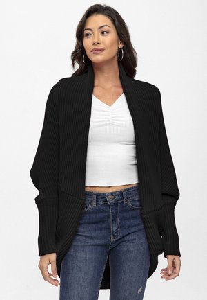 Jeune femme portant un cardigan noir oversize sur un haut blanc froncé et un jean bleu, debout devant un fond blanc uni.