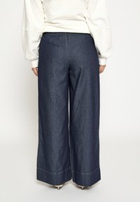 Široké rifľové nohavice s tmavým praním, elastickým pásom a kontrastným prešitím na leme. Hladká textúra s voľným strihom.