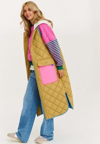 Prešívaná žltá bunda s veľkou ružovou vreckom, nosená cez pruhovanú ružovú a zelenú košeľu. Vidieť modré akcenty a svetlošedú mikinu.