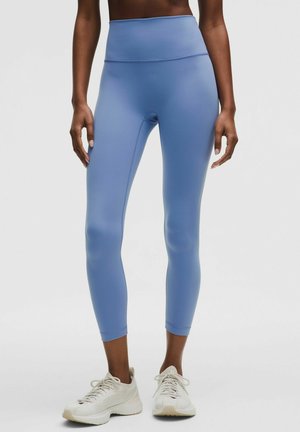 Frau trägt hellblaue Leggings mit hohem Bund und weiße Laufschuhe, steht vor einem einfarbigen Hintergrund.