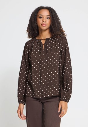 Femme aux cheveux bouclés portant un chemisier marron à manches longues avec des pois blancs et un pantalon marron assorti, debout sur fond blanc.
