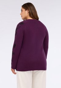 Lila stickad tröja med svarta blommönster på ärmarna, ribbade muddar och nederkant. Mjuk textur med avslappnad passform och rund halsringning.