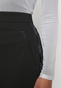 Černá sukně s úzkým páskem, která má boční kapsu se zipem a bílé vyšívané logo "Calvin Klein" podél bočního švu.