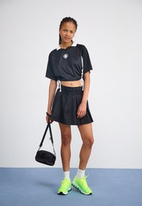 Top atletico nero corto con dettagli bianchi, abbinato a una gonna plissettata nera. Sneakers verde neon e una piccola borsa a tracolla nera completano il look.