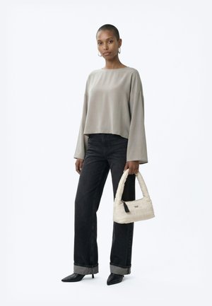 Femeie purtând un top gri larg cu mâneci lungi, blugi negri cu manșete, pantofi negri cu toc ascuțit, ținând în mână o geantă mică albă matlasată.