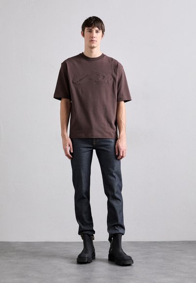 Camiseta de algodón marrón de manga corta con logotipo en relieve, combinada con jeans de mezclilla oscura y botines negros con cremalleras laterales.