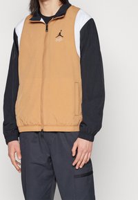 Vaaleanruskea vetoketjullinen takki, jossa mustia ja valkoisia yksityiskohtia. Takissa on joustavat hihansuut ja logo rinnassa. Pinta on sileä ja istuvuus rento.