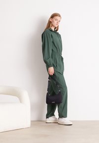 Tuta verde texurizzata con colletto e vita elasticizzata, abbinata a una piccola borsa nera Lacoste e sneakers bianche. Sfondo neutro.