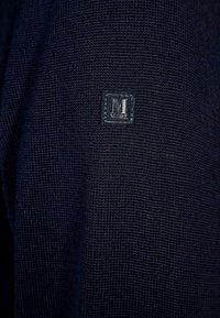Tumma sininen neulottu kangas, jonka keskellä on pieni suorakulmainen pala, jossa on jätetty tikattu "M MRZ" logo.