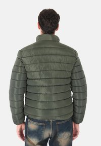 Giubbotto imbottito verde scuro, con cuciture orizzontali e colletto alto. Materiale liscio con una leggera lucentezza. Abbinato a jeans blu.