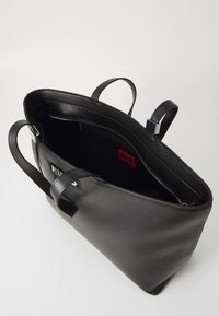 Czarna skórzana torba typu tote z dwoma uchwytami, wewnętrzną kieszenią na zamek i czerwoną metką „HUGO”. Charakteryzuje się gładką fakturą i usztywnionym kształtem.