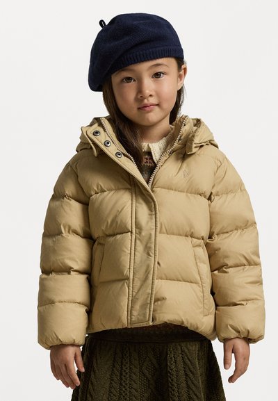 Beige dunjakke med hette, store kviltede paneler, knappelukking og en lyst brun genser under; båret med en mørk blå beret.