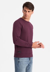 Sweatshirt bordeaux avec un col rond, des manches longues, des poignets côtelés et une finition texturée. Dispose d'une étiquette noire sur le côté.