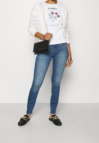 Hvid strikket cardigan over en floral grafisk t-shirt, parret med blå skinny jeans og sorte slip-on sko med guldfarvede detaljer. Sort crossbody taske.