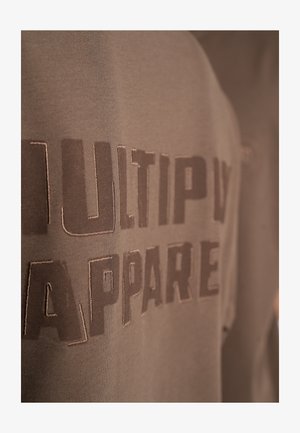 Ruskea puuvillaneule, jossa on suuri "MULTIPLY APPAREL" -kirjoitus. Kankaan pinta on sileä ja siinä on hienovarainen kiilto.