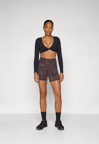 Schwarzes langärmeliges Crop-Top mit gedrehter Vorderseite, kombiniert mit braunen Leoprint-Hotpants mit hoher Taille und schwarzen Sportschuhen.