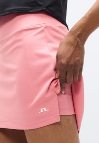 Ροζ αθλητική φούστα με ομαλή υφή, εφαρμοστή σχεδίαση και λευκή λεπτομέρεια λογότυπου. Ένα χέρι κρατά το ύφασμα στη ραφή στο πλάι.