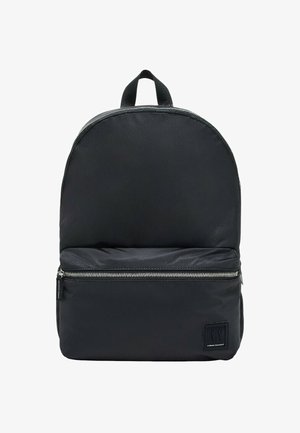 Mochila negra de nailon con forma redondeada en la parte superior, bolsillo frontal con cremallera y una etiqueta de tela en la parte inferior del frente. Cuenta con un asa superior para un transporte fácil.