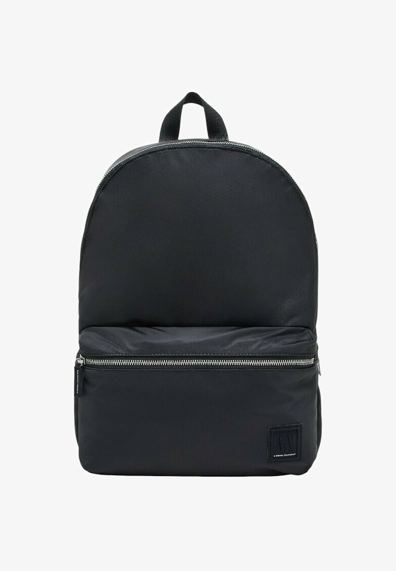 Zaino in nylon nero con parte superiore arrotondata, tasca frontale con zip e etichetta in tessuto nella parte bassa anteriore. Dotato di manico superiore per un facile trasporto.