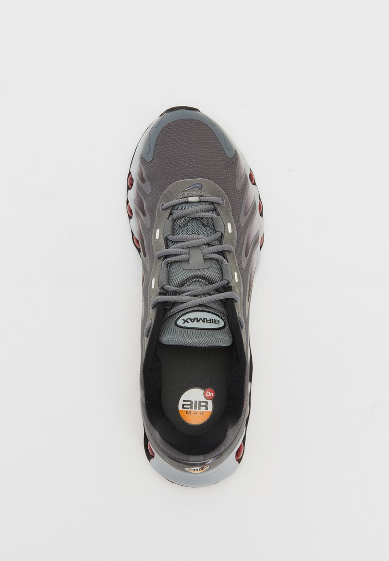 Zapatilla deportiva gris con parte superior de malla, diseño de cordones y amortiguación de aire visible. Detalles rojos a lo largo de la suela y marca en la plantilla.