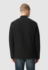 Czarna wełniana kurtka o strukturalnym kroju, z wysokim kołnierzem i prążkowanymi wykończeniami. Noszona na niebieskie dżinsy.