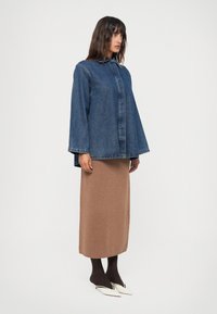 Camisa de ganga com colarinho e mangas largas, combinada com uma saia maxi de malha castanha e collants escuros. Sapatos brancos de bico fino completam o look.