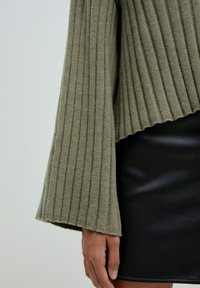 Olivengrüner gerippter Pullover mit weiten Ärmeln, übereinandergetragen über einem schwarzen Leder-Minikleid. Der Stoff erscheint texturiert mit sichtbaren Rippen.