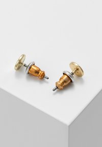 Auksiniai ir sidabriniai metaliniai smeigės su apvaliu viršumi ir tekstūruota, burbuliška baze su smeigtuku tvirtinimui, išdėstyti ant baltos plokštumos.