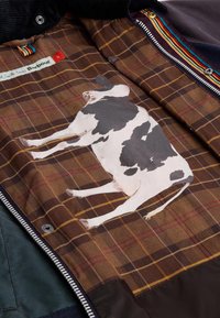 Barna kockás belső bélés, tehén illusztrációval. Cipzáras záródás, kontrasztos szövetbetétek és színes zsinór díszítéssel rendelkezik.